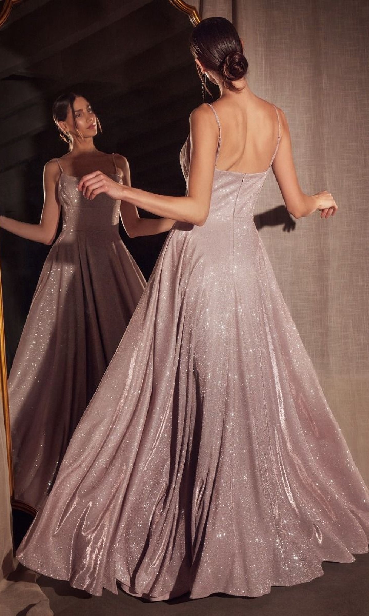 A woman stands before a mirror in the Classic Long Glitter Prom Dress: Amoris T213, featuring thin straps, an open back, and a flared sparkling floor-length skirt.