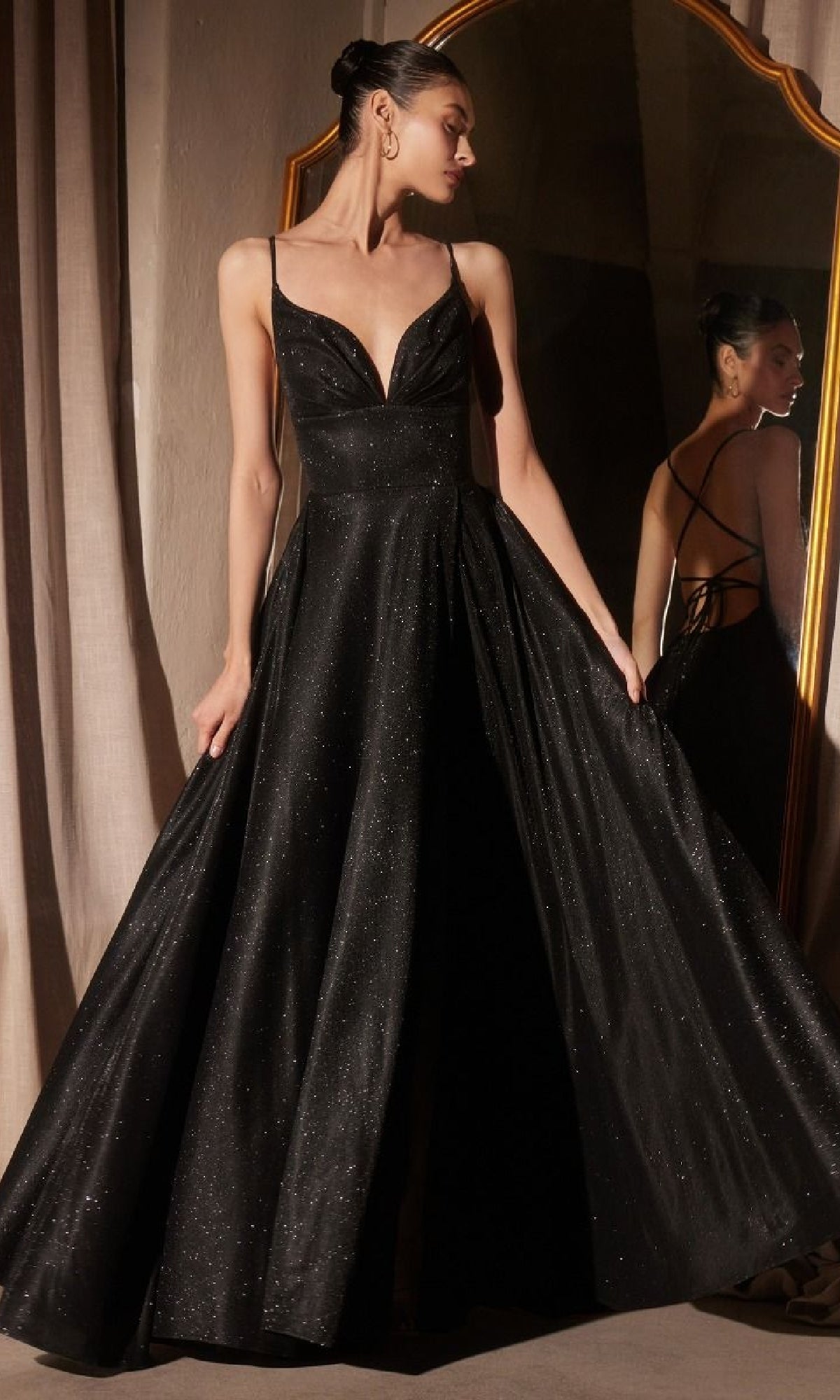 A woman stands in front of a mirror wearing the Amoris T214 Strappy-Back Glitter Prom Gown—a floor-length, sleeveless black dress with a deep V-neck and subtle shimmer.
