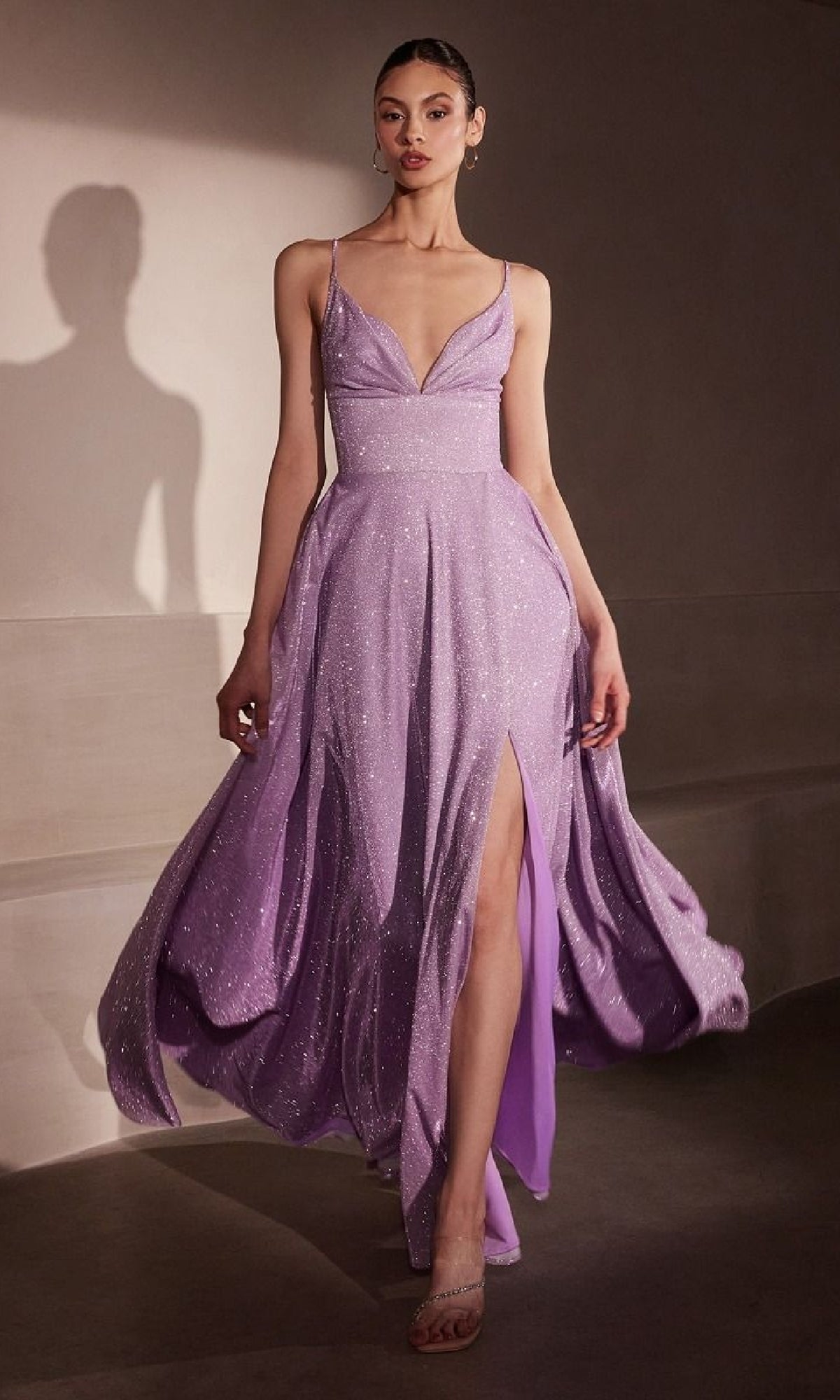 A woman walks indoors in the Amoris T214 Strappy-Back Glitter Prom Gown—lavender, shimmery, with thin straps, a deep neckline, and a high slit.