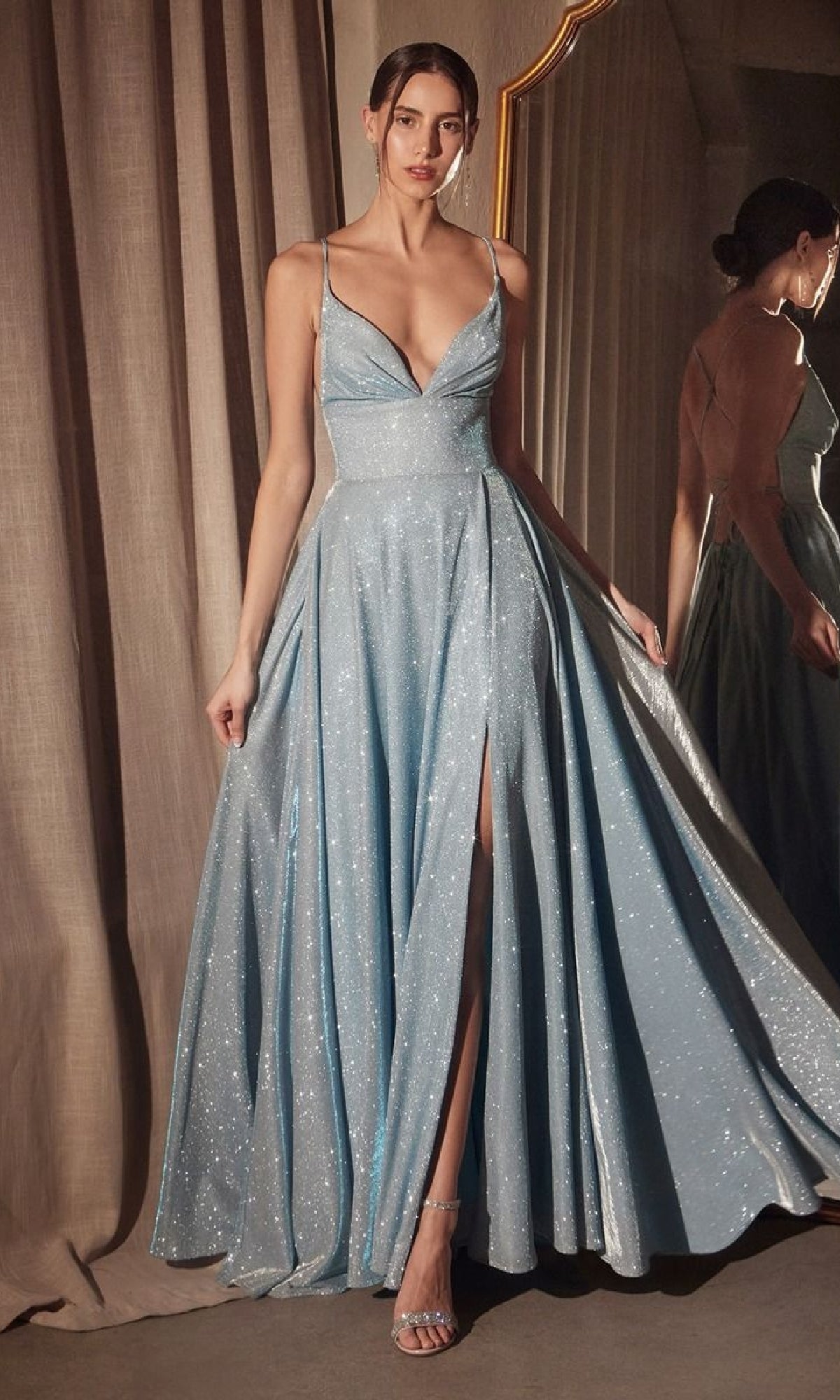 A woman in the Strappy-Back Glitter Prom Gown: Amoris T214, featuring a light blue shimmer, thin straps, deep neckline, high slit, and open back, stands by a mirror and curtain.