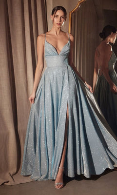 A woman in the Strappy-Back Glitter Prom Gown: Amoris T214, featuring a light blue shimmer, thin straps, deep neckline, high slit, and open back, stands by a mirror and curtain.