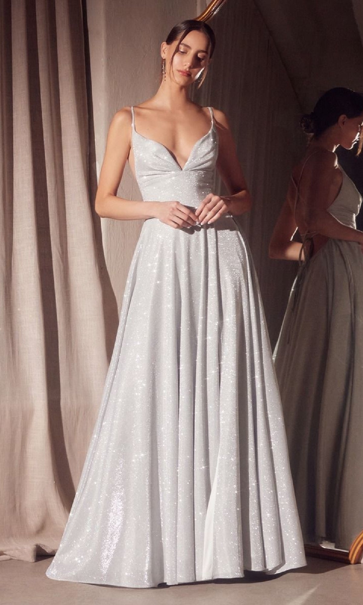 A woman stands indoors wearing the Strappy-Back Glitter Prom Gown: Amoris T214, a floor-length, sleeveless silver dress with a deep V-neckline, reflected in a nearby mirror.
