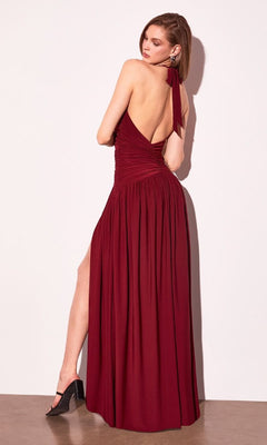 A woman in the Amoris T502 Backless Halter Prom Dress with Slit by Cinderella Divine, a sleeveless burgundy gown, stands against a white wall and looks over her shoulder.