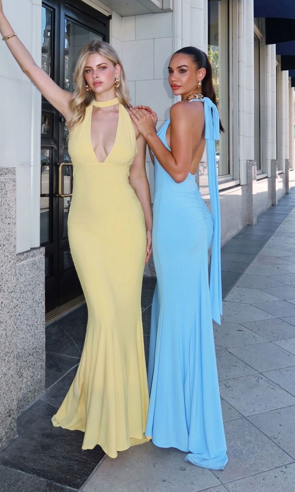 Two women pose confidently on a city sidewalk, each wearing the Astee T505 Long Prom Dress—one in yellow, one in light blue—crafted from form-fitting stretch fabric as they stand side by side in front of a building.