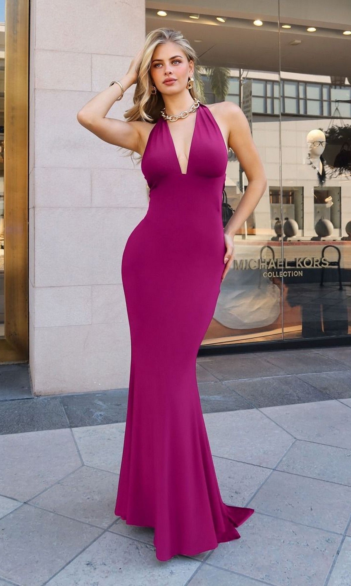 A woman poses outside a Michael Kors store, wearing the Astee T505 Long Prom Dress in orchid—a form-fitting gown with a deep V neckline—accessorized with a gold necklace and bracelet, her hand stylishly placed in her hair.