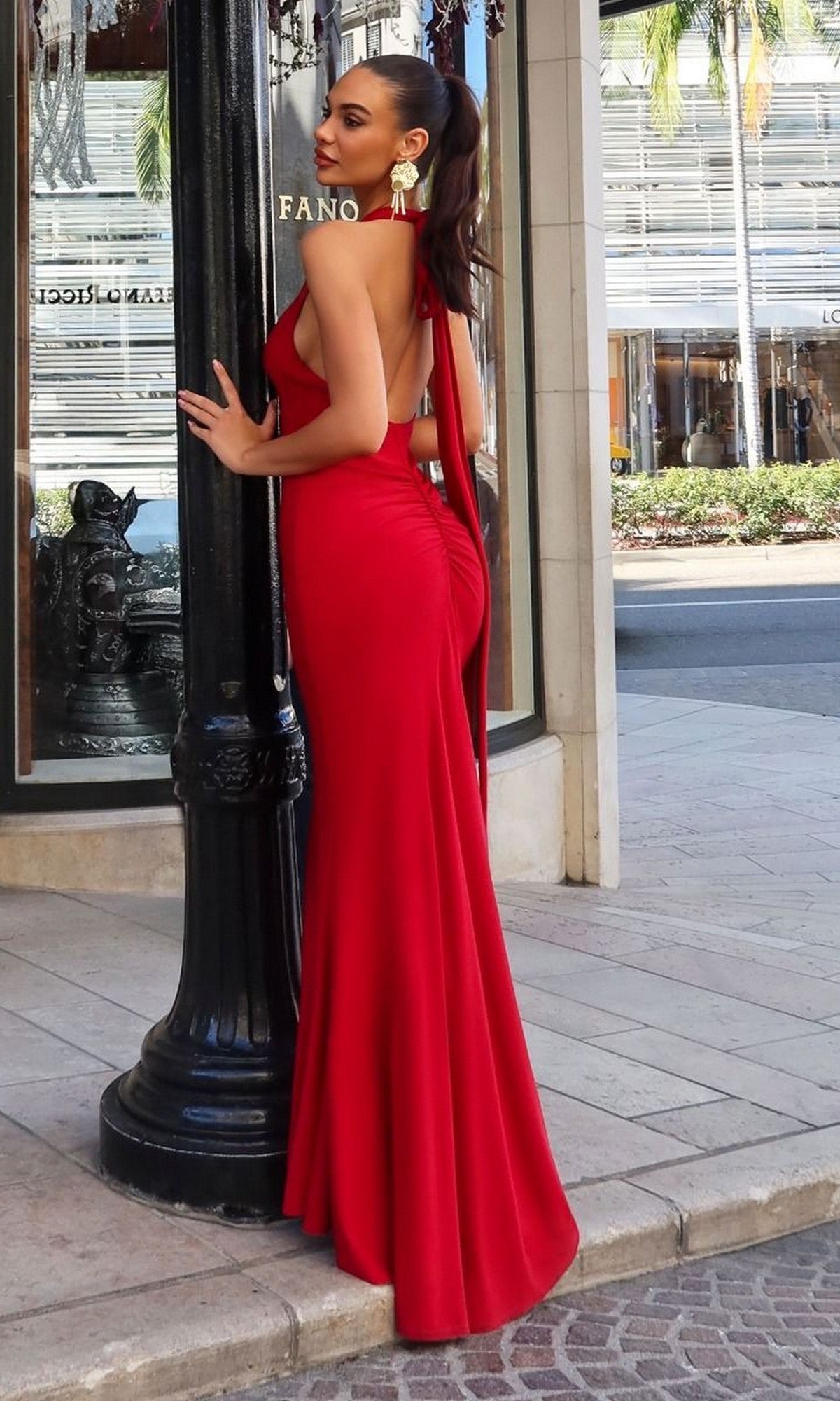 A woman wearing the Long Prom Dress: Astee T505, a red form-fitting halter gown in stretch fabric, stands outdoors by a black lamppost on a stone sidewalk with storefronts and palm trees behind her.