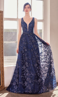 A woman stands in front of large windows wearing the Amoris T606 Embroidered-Lace Long Prom Dress, a sleeveless, deep V-neck navy gown with floral lace and sequin embellishments.