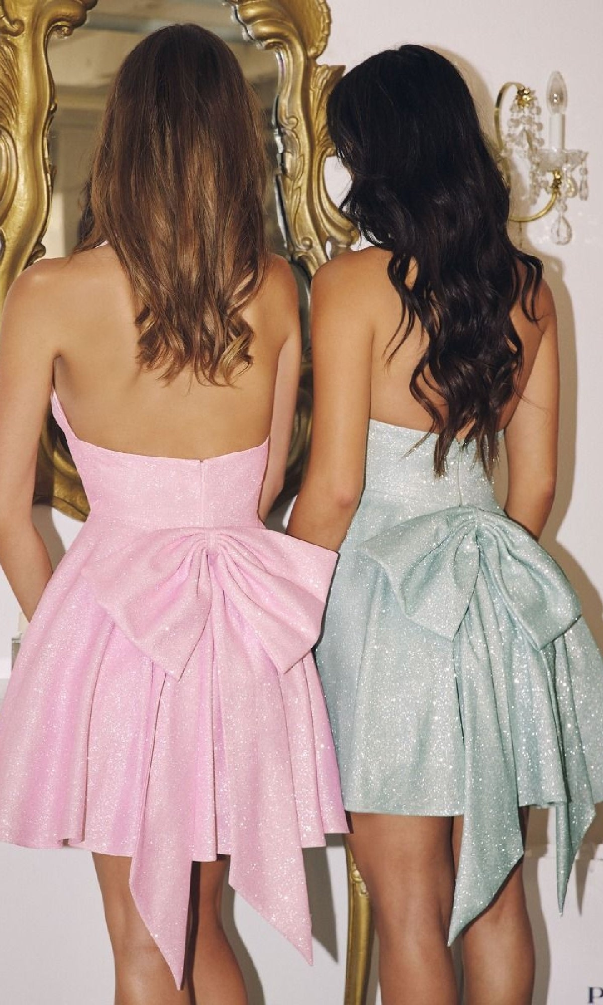 Two women wearing pink and green dresses with bows on the back standing in front of a decorative mirror.