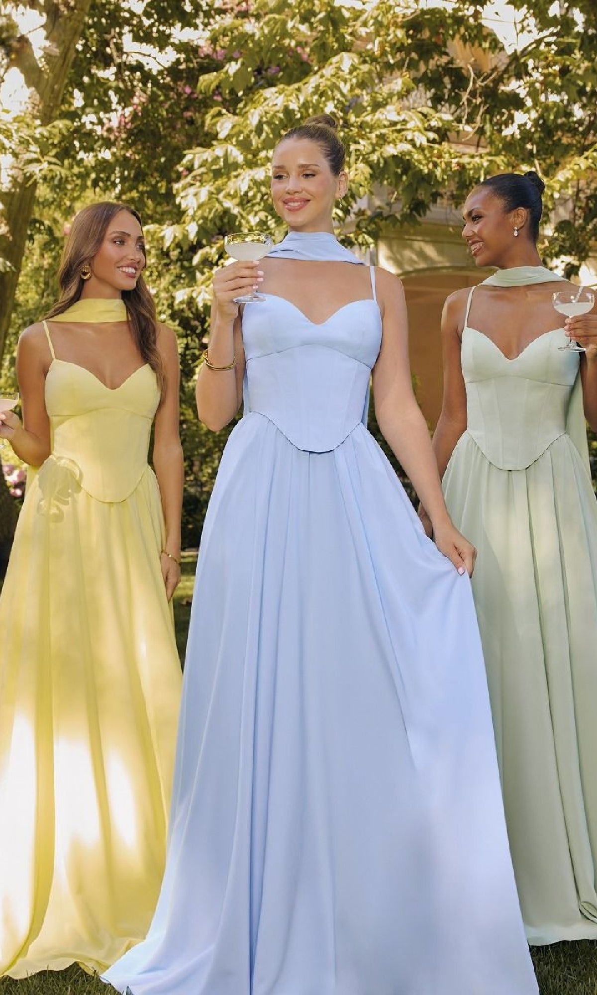 Three women in formal dresses standing outdoors with trees in the background