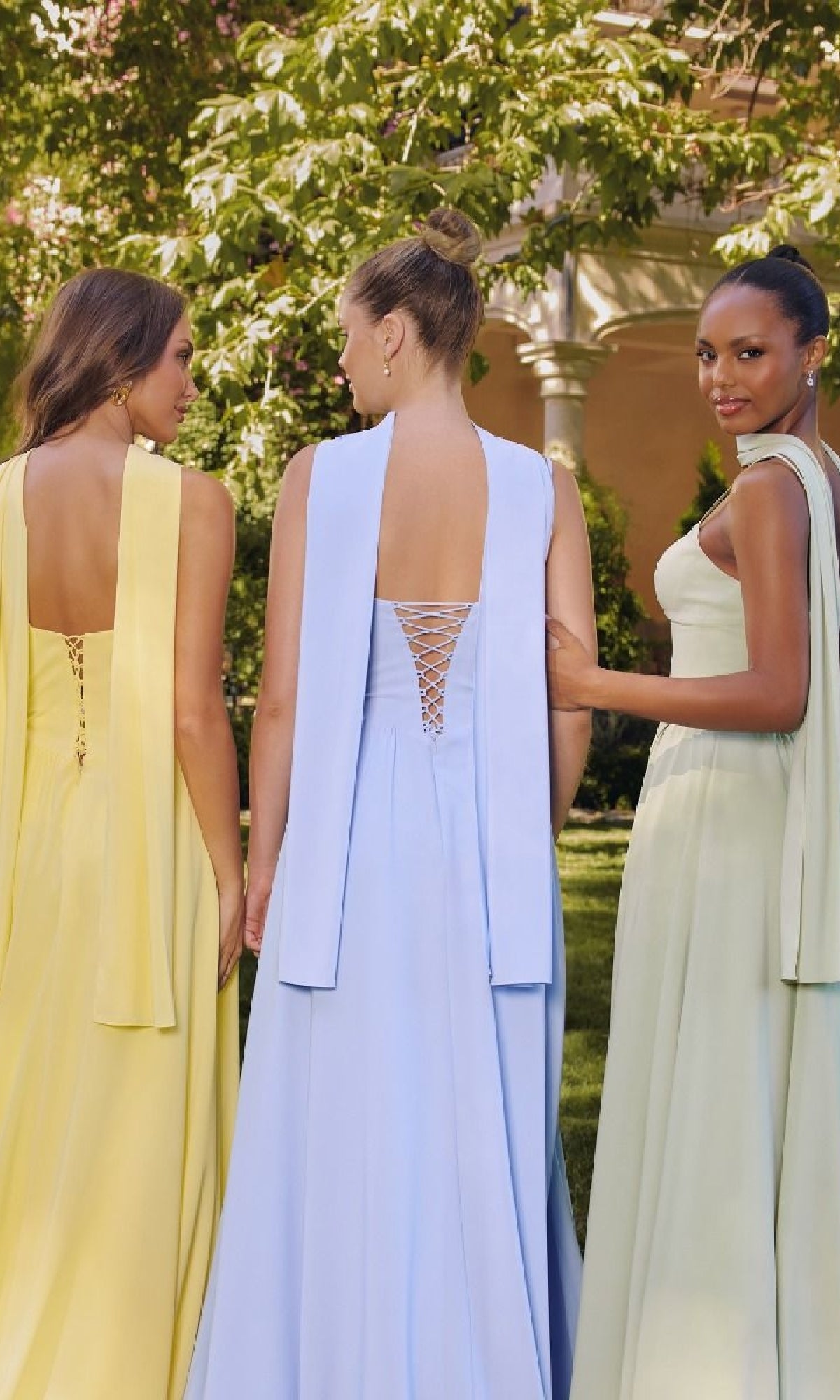 Three women wear Amoris T711 Lace-Up Long Bridesmaid Dresses with draped panels outdoors; two show off the lace-up backs, while one turns and smiles at the camera.