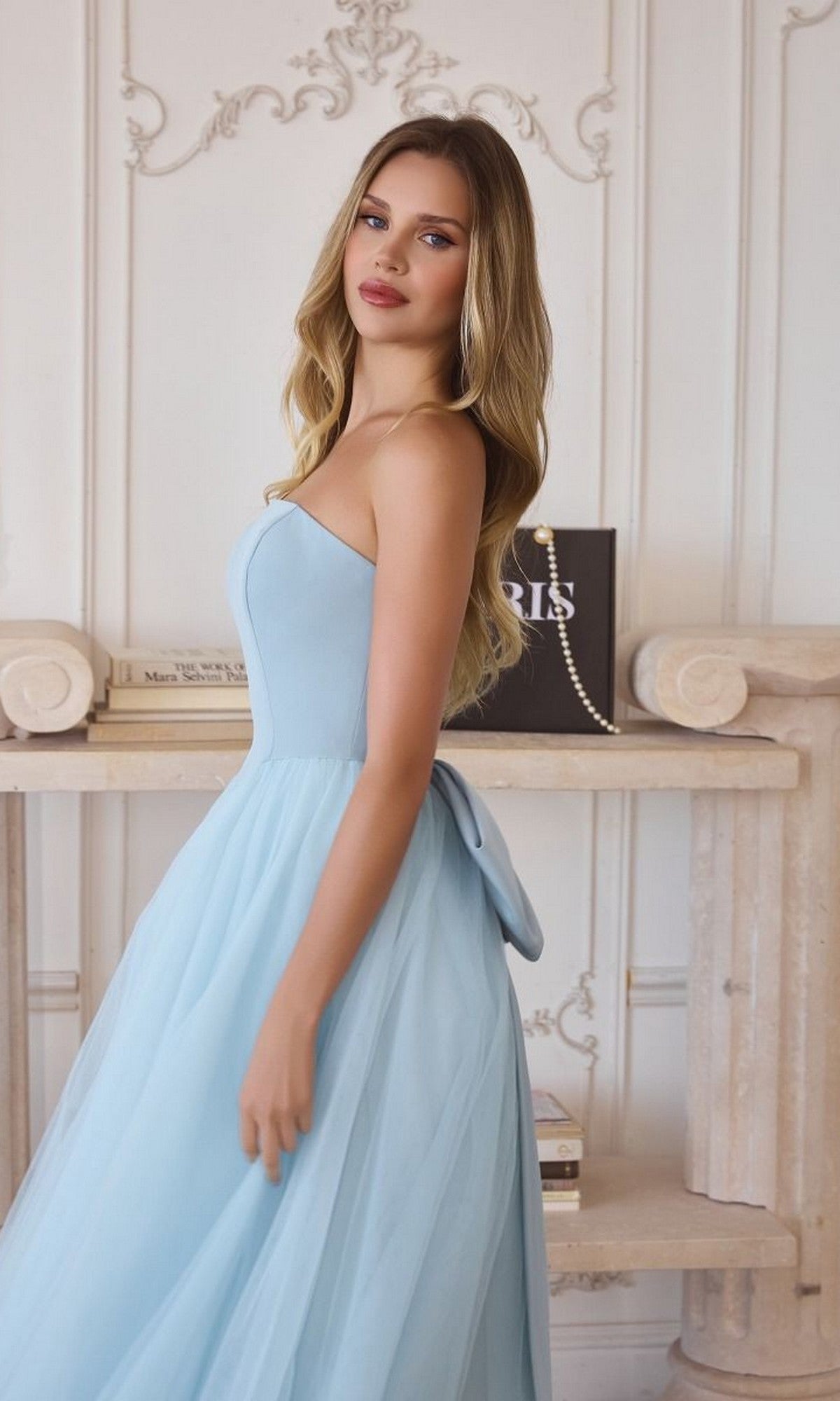 A woman in an affordable, formal setting showcases the Classic Strapless Prom Ball Gown: Amoris T712—a light blue gown—while standing sideways in an elegant room with white walls and a decorative bookshelf.