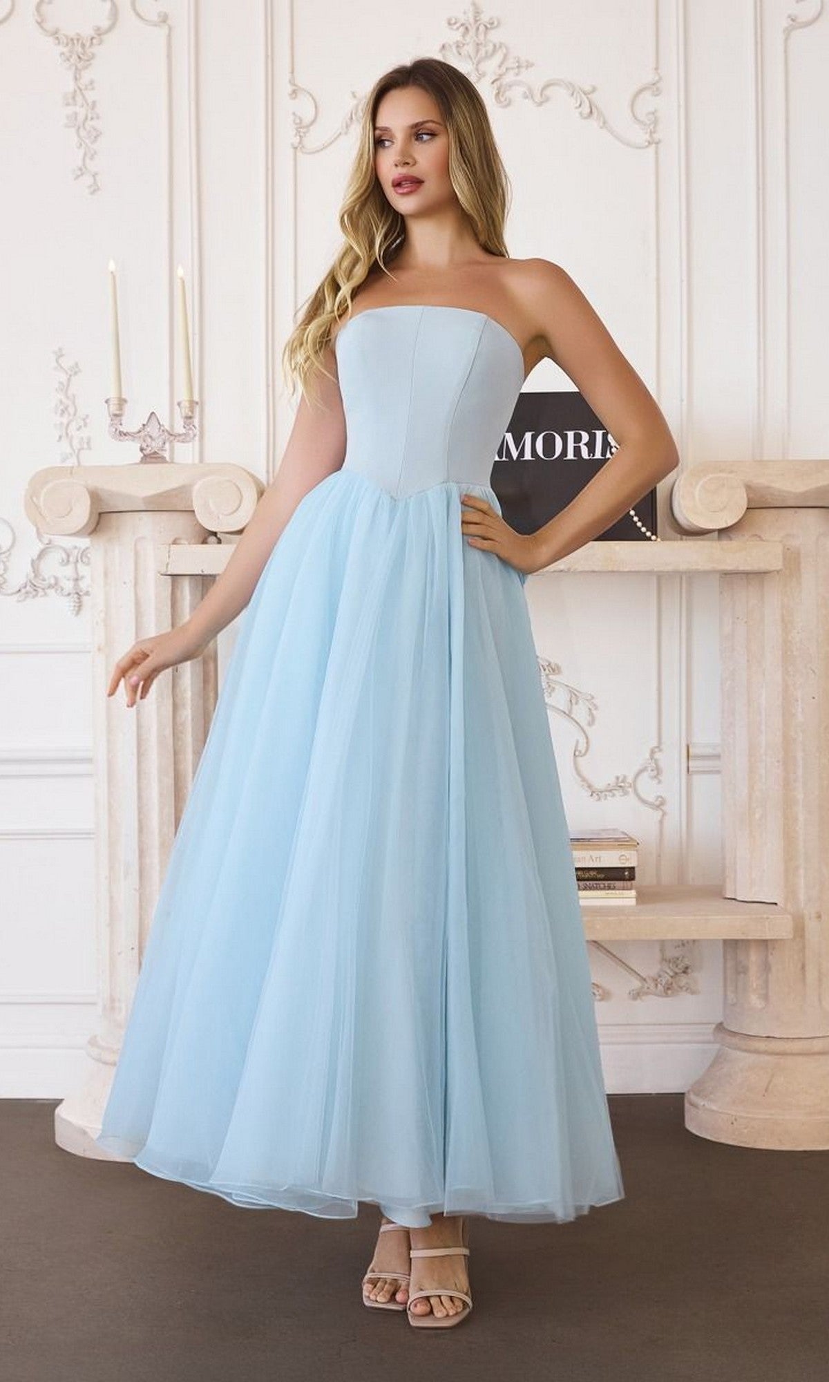 A woman in the Classic Strapless Prom Ball Gown: Amoris T712, a light blue long dress, poses indoors by a mantelpiece with one hand on her hip.