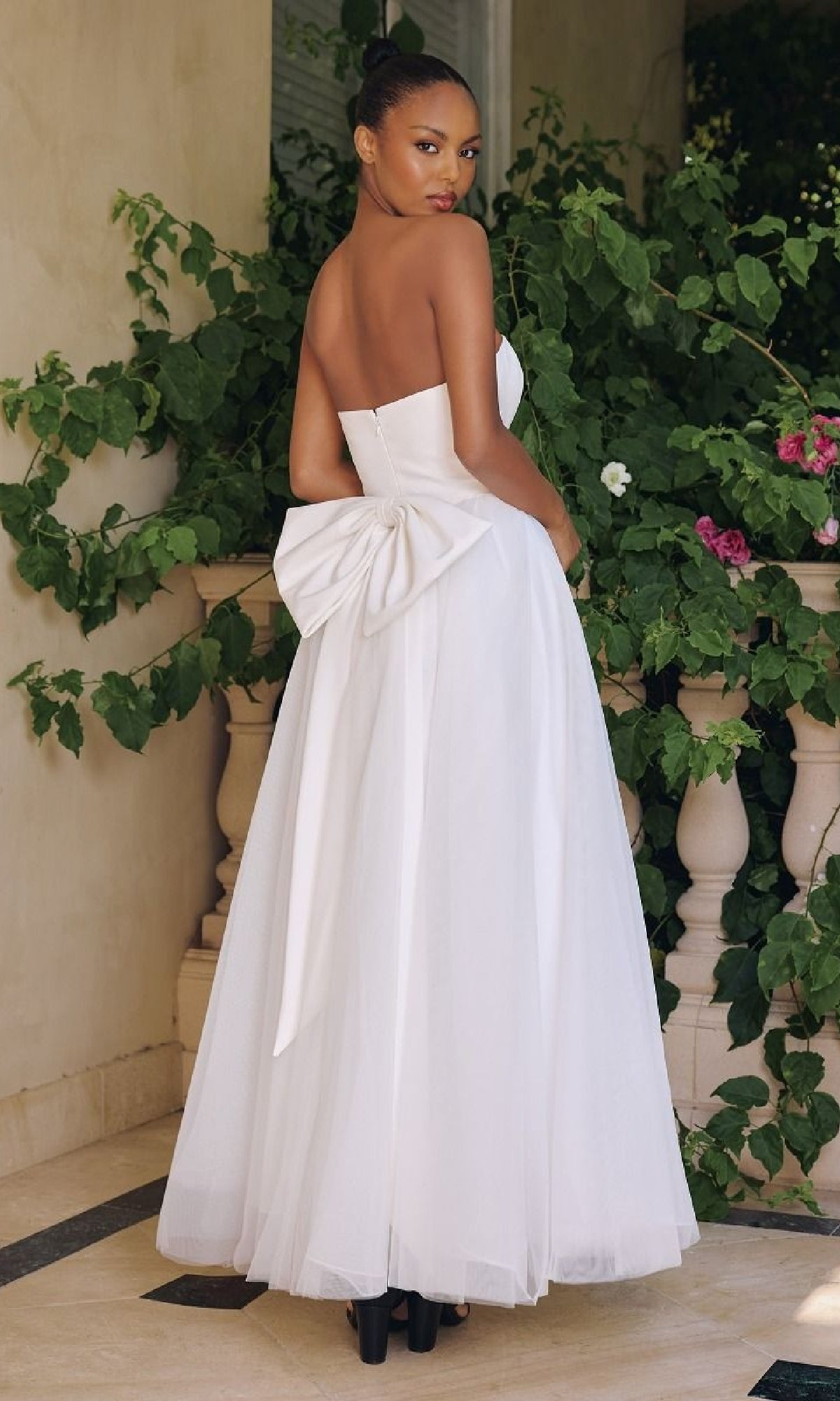 Woman in a white strapless gown with a large bow at the back, standing outdoors with greenery.
