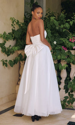 Woman in a white strapless gown with a large bow at the back, standing outdoors with greenery.