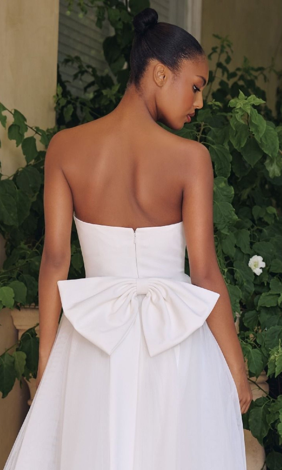 Woman wearing a white strapless gown with a large bow detail, standing outdoors with greenery in the background.
