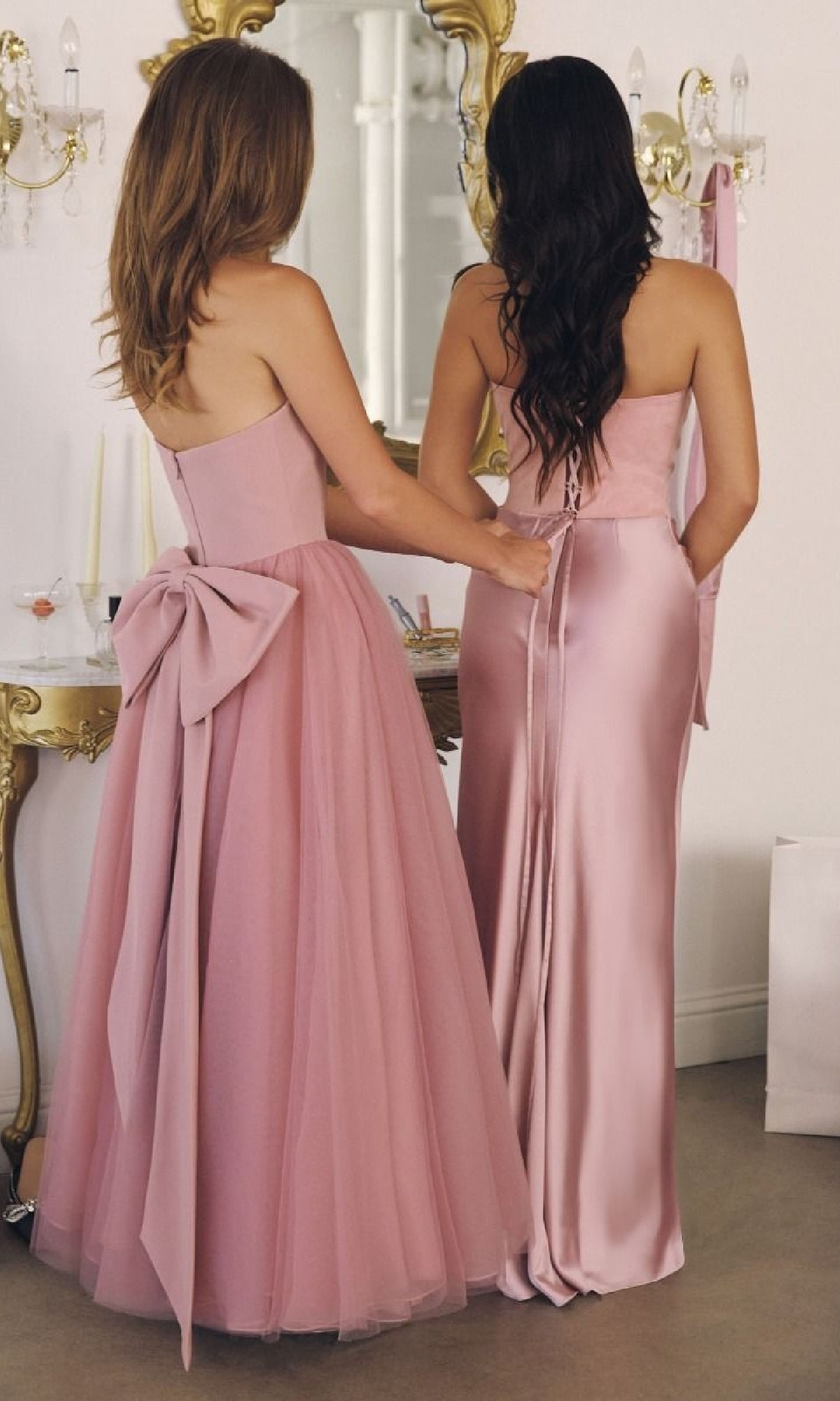 Two women in Classic Strapless Prom Ball Gown: Amoris T712 pose by a mirror; one shows off a tulle style with a statement bow, while the other stuns in a fitted satin look.