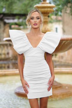 Woman wearing the Velvi Ruffle-Shoulder Short Party Dress Tess—a white, ruched mini dress with bold sleeves—stands before a blurred outdoor fountain and greenery.