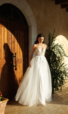 A woman in the Tina Holly TW480 long formal dress with a strapless white bodice and tulle A-line skirt stands outside by a wooden door, sunlight casting gentle shadows on the wall and floor.