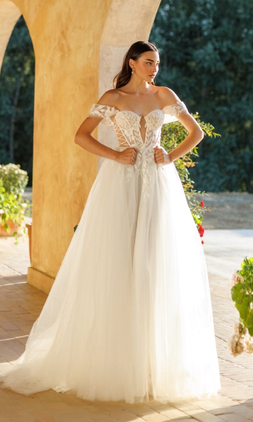 A woman wearing the Tina Holly TW480 long formal dress, featuring an off-the-shoulder corset bodice and tulle A-line skirt, stands outdoors by arches and lush greenery.