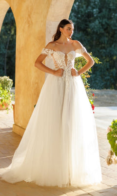 A woman wearing the Tina Holly TW480 long formal dress, featuring an off-the-shoulder corset bodice and tulle A-line skirt, stands outdoors by arches and lush greenery.
