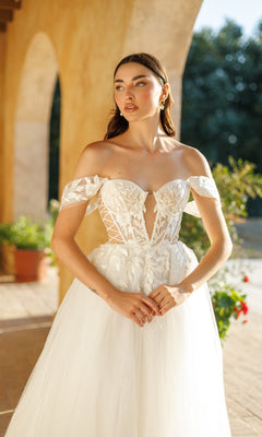A woman stands outdoors in natural light, wearing the Long Formal Dress: Tina Holly TW480, an off-shoulder white bridal gown with a corset bodice and lace floral appliqué details.