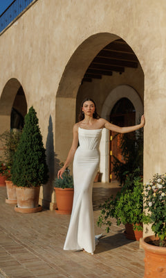 A woman stands under an arched walkway by potted plants on a sunny day, wearing the Off-White Long Bridal Gown: Tina Holly TW485—an elegant, strapless, floor-length dress flowing gracefully.