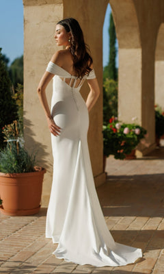A woman in the Off-White Long Bridal Gown: Tina Holly TW485 stands outdoors on a sunlit patio, framed by stone columns and potted plants.