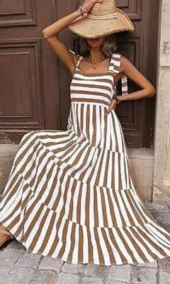 A woman poses on stone pavement in front of a wooden door, wearing the Striped Casual Relaxed-Fit Maxi Dress and a wide-brimmed straw hat, with one arm raised to her head.
