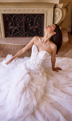 A woman wearing the Long Wedding Dress: Andrea and Leo WL016, featuring a strapless bodice and ruffle tulle a-line skirt, sits on the floor before an ornate fireplace with her head tilted upward and eyes closed.