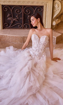 A woman wearing the Andrea and Leo WL016 long wedding dress, featuring a strapless lace-applique corset bodice and ruffled tulle A-line skirt, sits on a stone floor before an ornate fireplace.