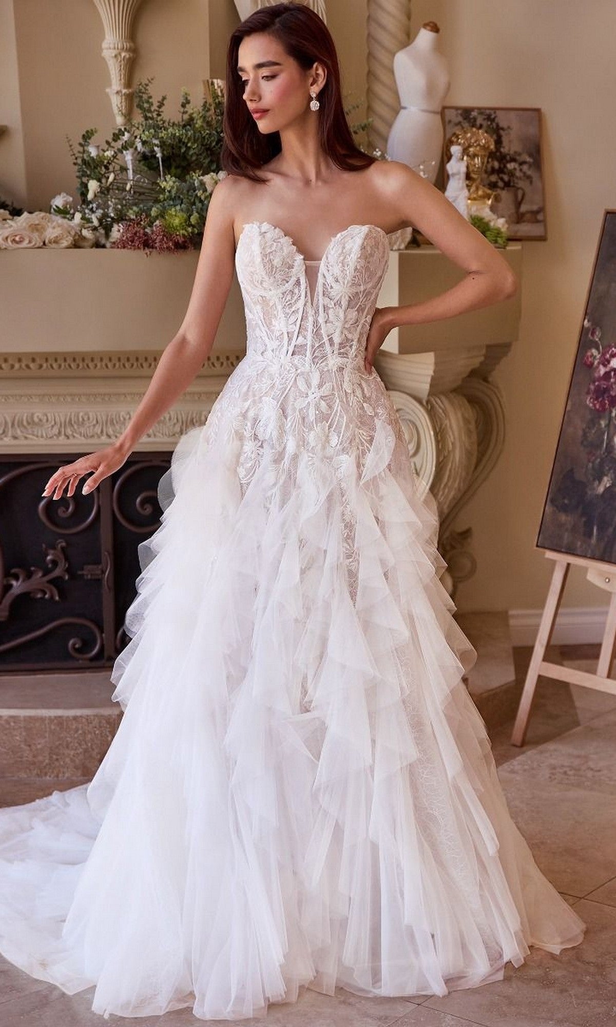 A woman wearing the Long Wedding Dress: Andrea and Leo WL016, featuring a strapless lace-applique corset bodice and ruffle tulle A-line skirt, stands indoors by a fireplace with floral decorations.