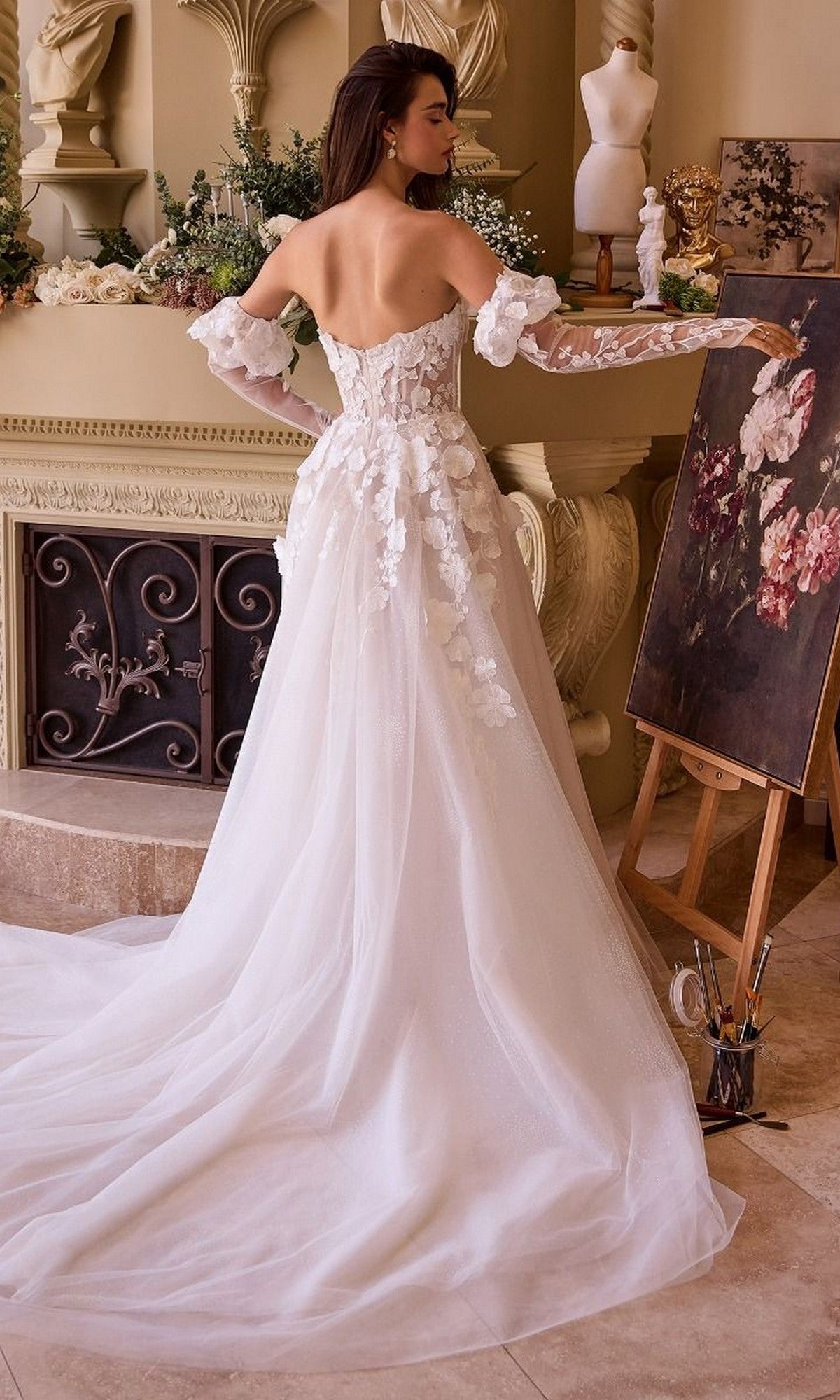 A woman in the Andrea and Leo WL017 long wedding dress with 3D floral appliques stands by a fireplace, painting flowers on a canvas in an elegant room.