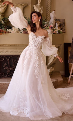 A woman poses indoors before floral decorations, wearing the Andrea and Leo WL018 long wedding dress featuring a white embroidered design, sweetheart neckline, off-shoulder style, and sheer puffy sleeves.