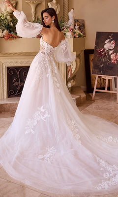 A woman wearing the Andrea and Leo WL018 Long Wedding Dress, featuring a strapless off-shoulder lace design with floral details and a long train, stands indoors near a fireplace and elegant floral decorations.