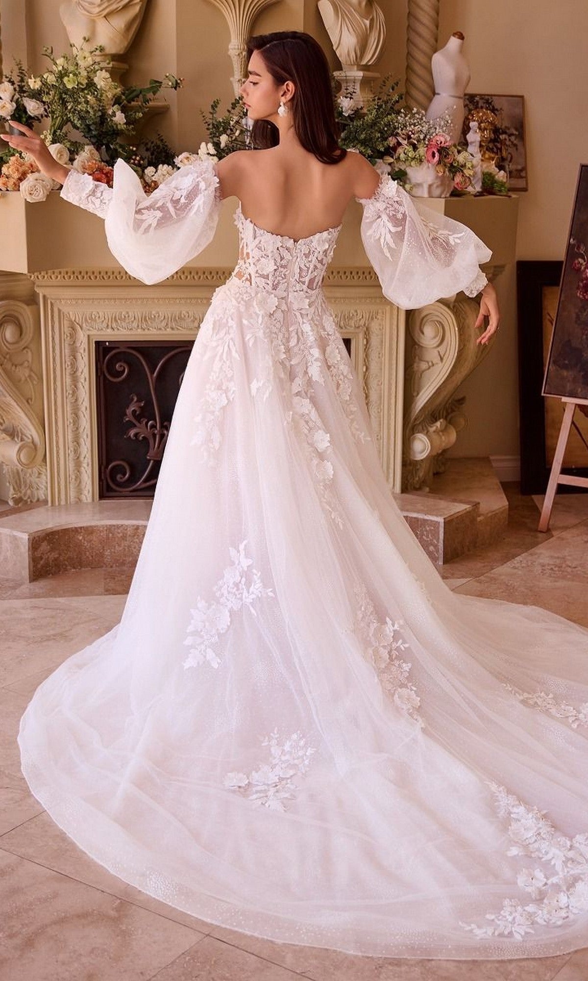 A woman in the Andrea and Leo WL018 long wedding dress, featuring an off-the-shoulder sweetheart neckline, lace embroidery, and a long train, stands indoors facing an ornate fireplace.