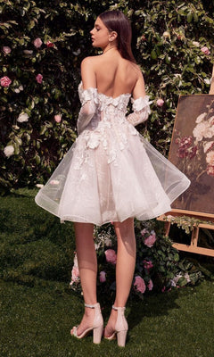 A woman stands outdoors in the Andrea and Leo WL020 Short White Party Dress with floral appliques and sheer sleeves, surrounded by greenery, flowers, and a nearby floral painting.