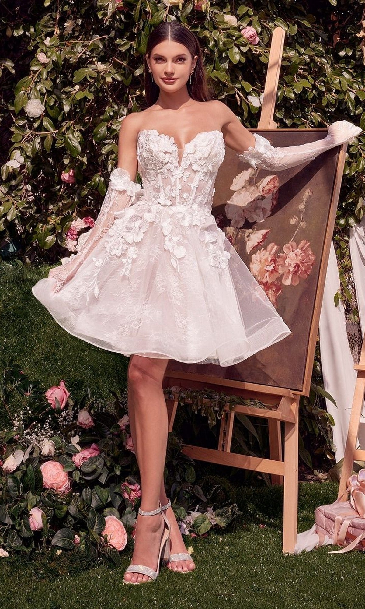A woman in the Andrea and Leo WL020 short white party dress poses outdoors beside an easel and flowers.