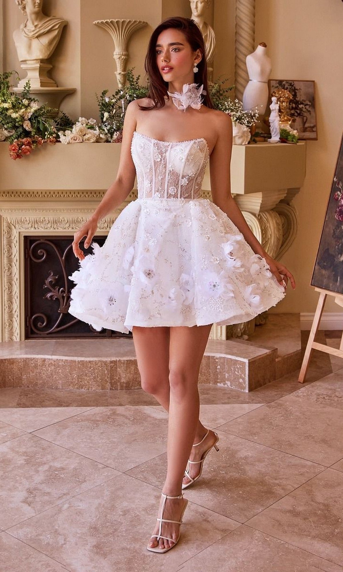 A woman wears the Short White Dress: Andrea and Leo WL025, featuring 3D floral appliques and a sheer bodice, while standing indoors amid classical decor and lush floral arrangements.