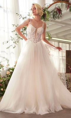 A woman stands indoors near flowers and a mirror, holding part of her skirt while wearing the Andrea and Leo WL029 long wedding dress—a sleeveless gown with a lace corset bodice and an A-line tulle skirt.