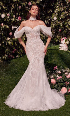 A woman in the Andrea and Leo WL031 long wedding dress in off-white, featuring a strapless lace mermaid design with sheer sleeves and a removable organza shawl, stands in a blooming garden.
