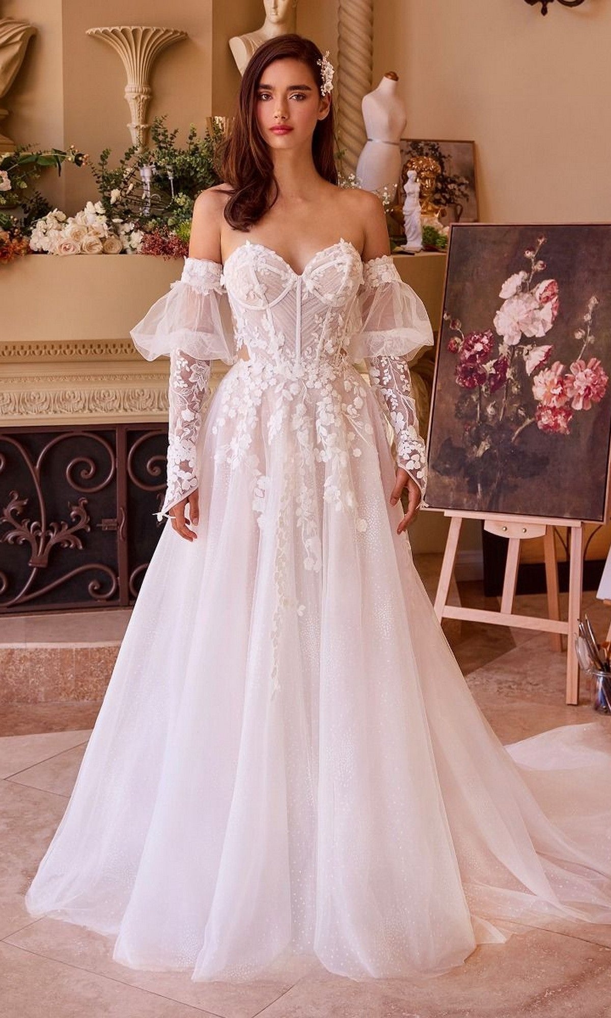 A woman in the Long Wedding Dress: Andrea and Leo WL032, featuring a strapless sweetheart neckline and white lace floral details, stands indoors beside a floral painting and decorative plants.