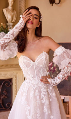 A woman wearing the Andrea and Leo WL032 long wedding dress with a strapless sweetheart neckline and floral lace details poses indoors, her gloved hand partially covering her face.