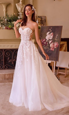 A woman in the Andrea and Leo WL032 long strapless wedding dress with floral appliqués and an elegant A-line skirt stands indoors near a floral painting and an easel.