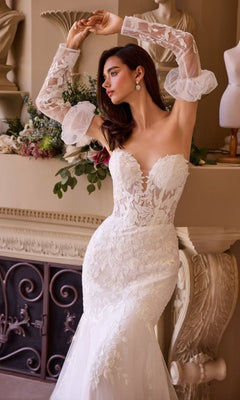 A woman poses indoors near a decorated fireplace, wearing the Andrea and Leo WL033 long wedding dress with strapless neckline, sheer ruffled sleeves, and delicate floral lace appliques.