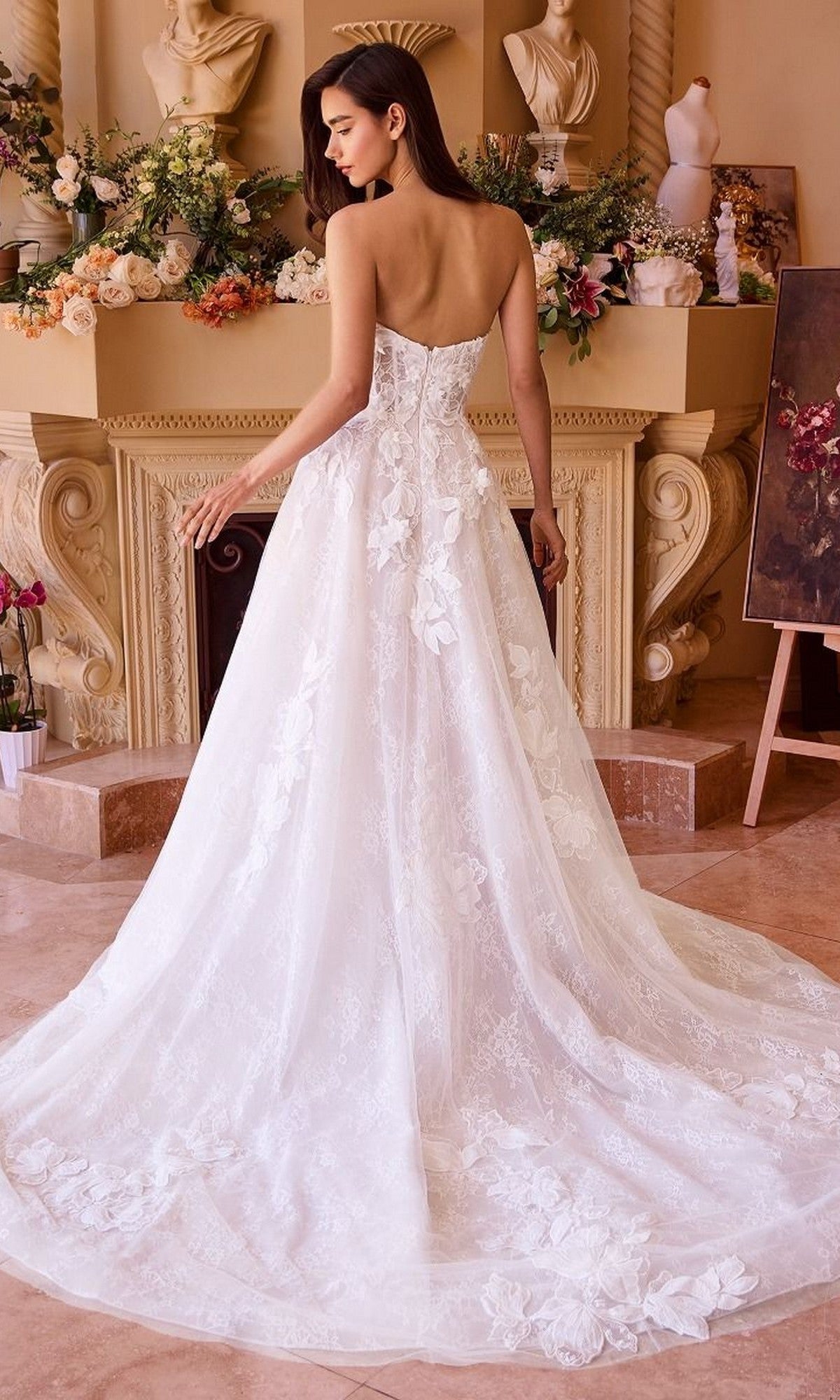 A woman in the Andrea and Leo WL034 long wedding dress, featuring a strapless design with floral lace appliques and a long train, stands indoors facing away, surrounded by flowers and classical decor.