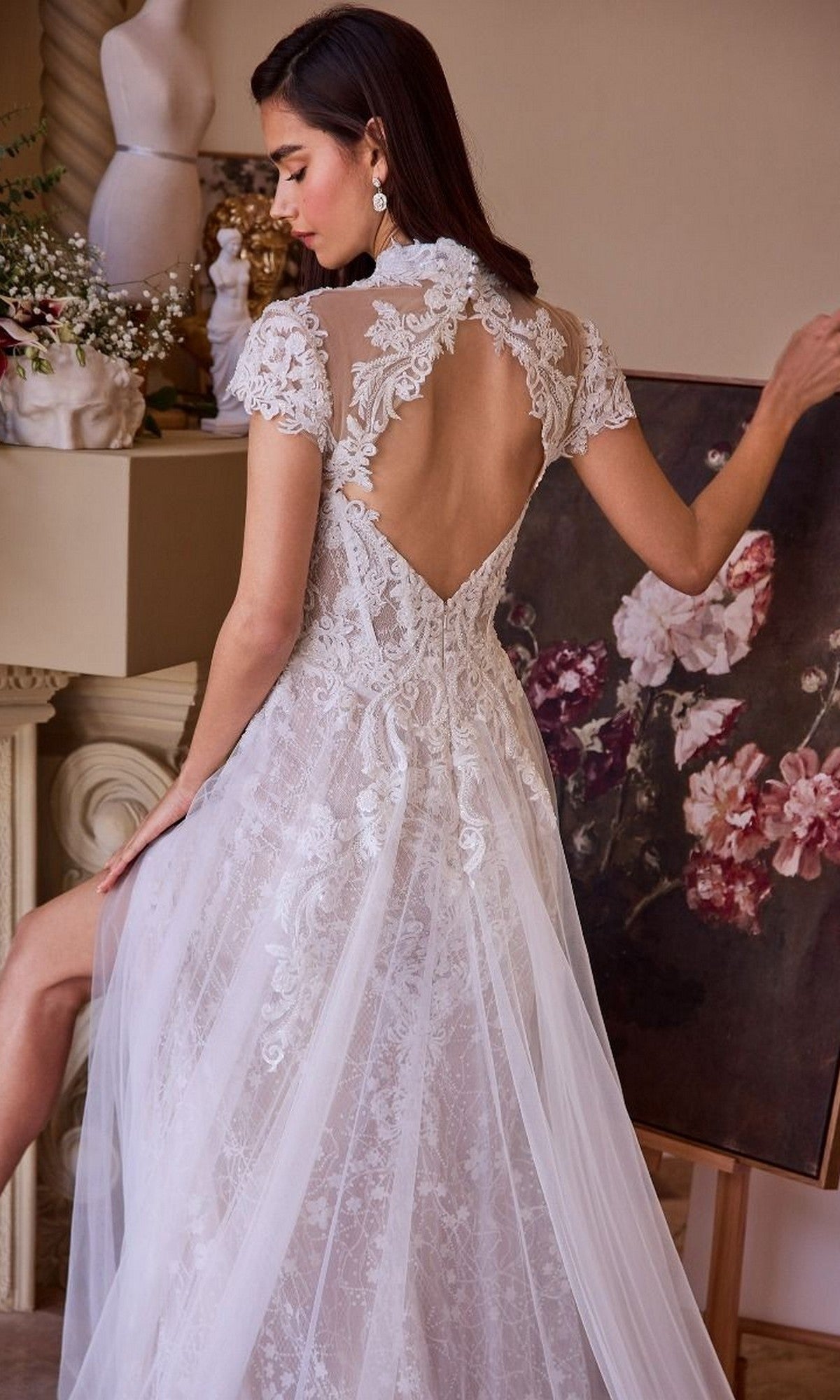A woman wears the Long Wedding Dress: Andrea and Leo WL035, featuring a sheer back and floral embroidery, as she stands near a floral painting.