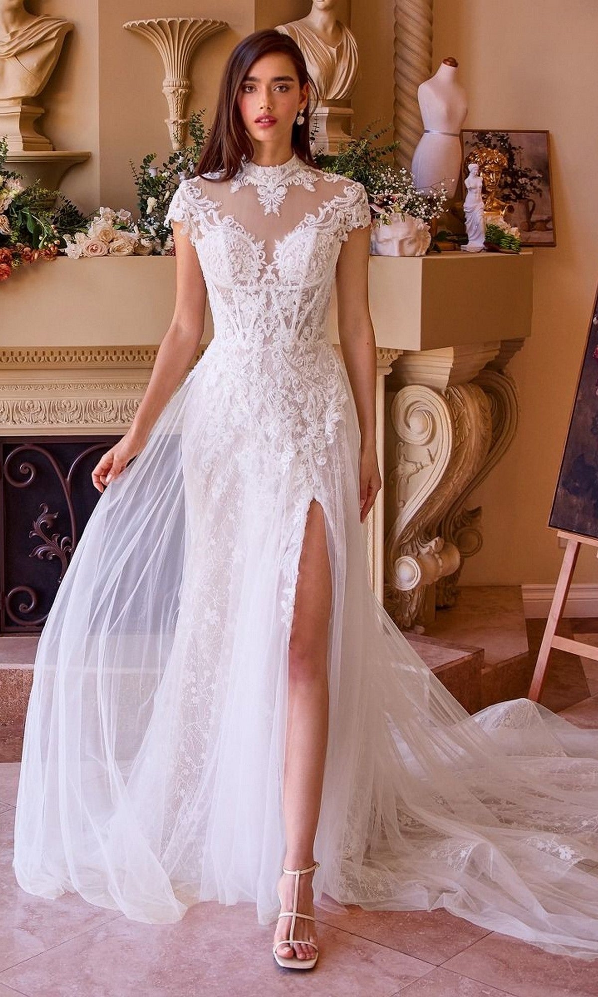 A woman wearing the Andrea and Leo WL035 long wedding dress, a white lace gown with a high slit and sheer neckline, stands indoors by a decorated mantel.