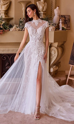 A woman stands indoors near a decorated fireplace, showcasing the Andrea and Leo WL035 long wedding dress with a high neckline, cap sleeves, and a thigh-high slit for an elegant bridal look.