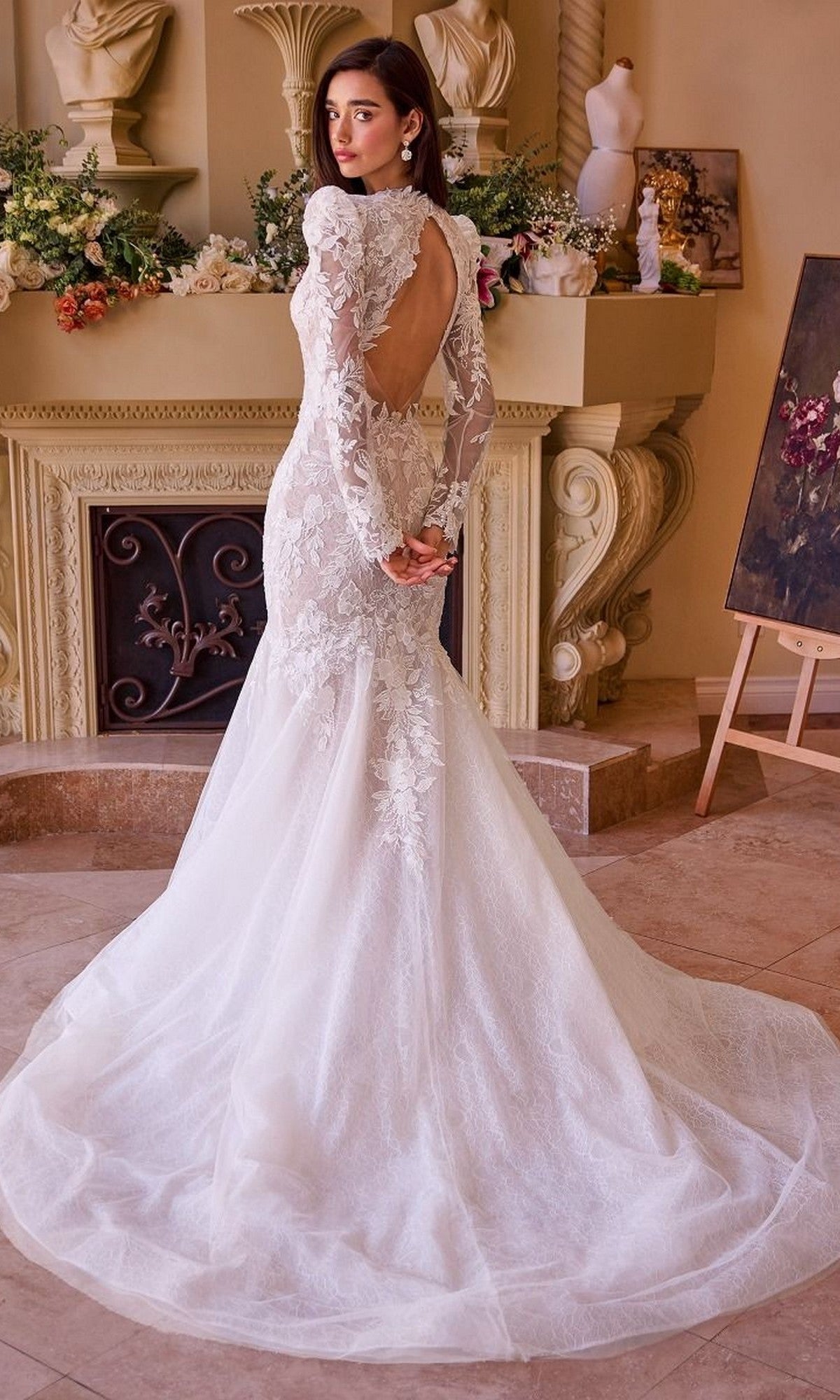 A woman wearing the Andrea and Leo WL036 long wedding dress, featuring lace, long sleeves, a mermaid silhouette, and an open back, stands indoors before a decorated mantelpiece, gazing over her shoulder.