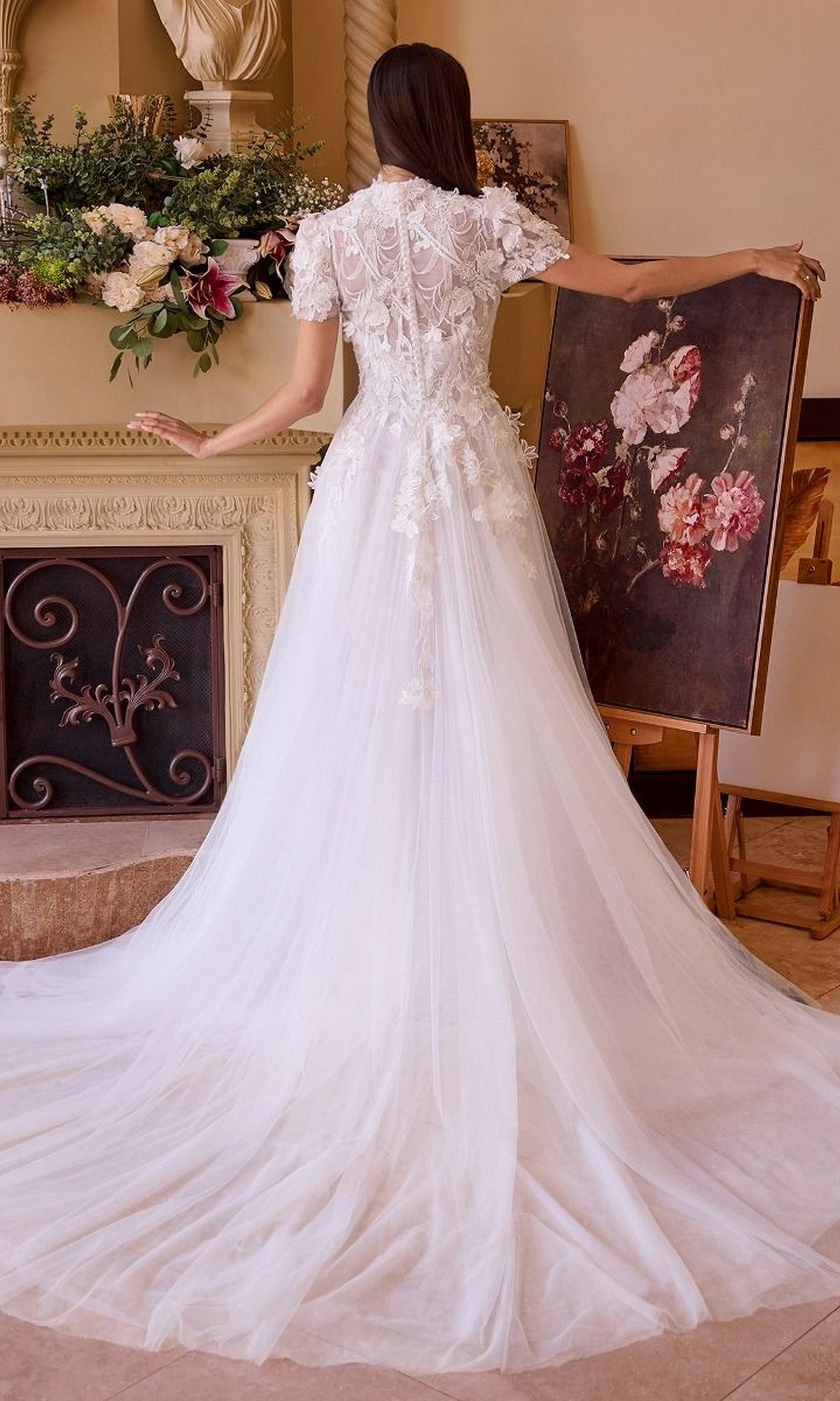 A woman wearing the Andrea and Leo WL040 long wedding dress with a sweetheart neckline stands with her back to the camera, holding a floral painting near a decorated fireplace and floral arrangement.